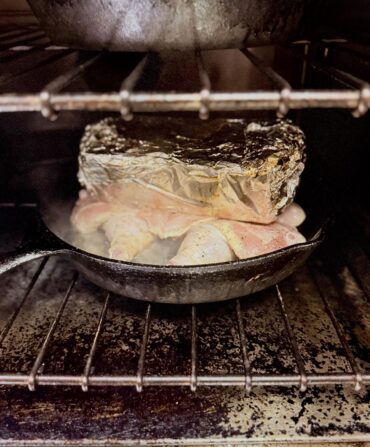 A flattened chicken under a tinfoiled wrapped brick in a cast iron pan in an oven