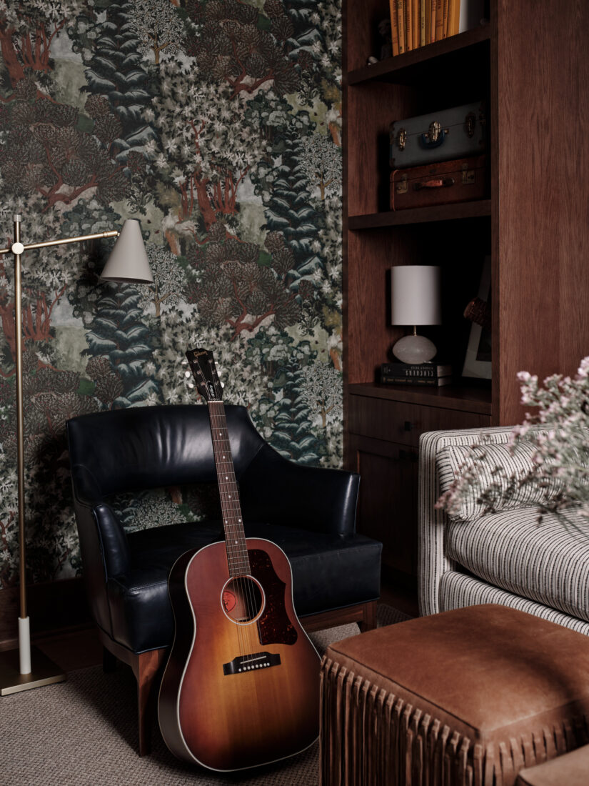 A guitar stands against a leather chair in a room with floral wallpaper