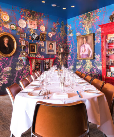 Inside a colorful dining room with floral wallpaper and eclectic artwork on the wall. A set table sits in the middle