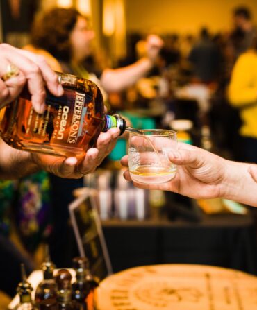 A bottle pours bourbon into a glass cup a person is holding.