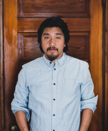 An asian man with a beard wearing a blue button up shirt standing in front of a wood door.