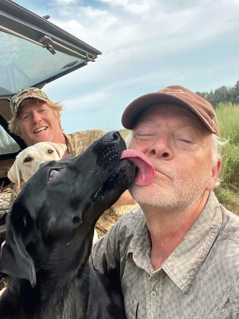 Two men with their dogs. One dog is licking one man's face.