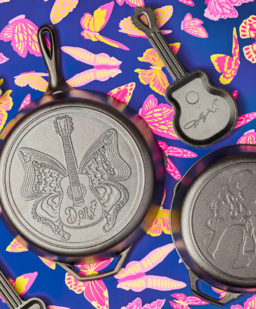 Four cast iron pans with etchings of butterflies, Dolly Parton's head and signature, and a guitar-shaped pan. They are on a blue, pink, and yellow butterfly-patterned background