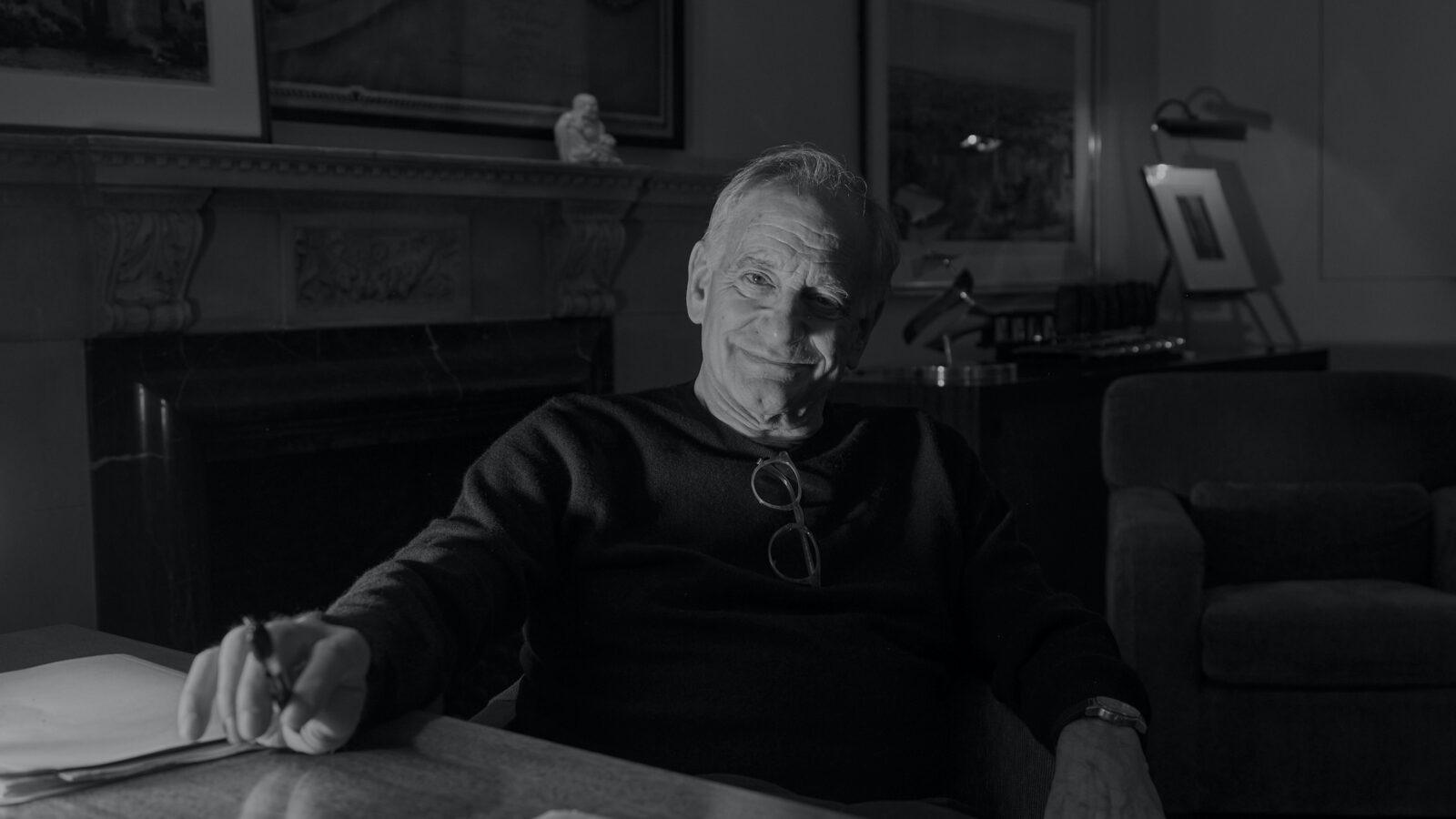 A black and white photo of a man sitting at a writing desk with a pen in his hand and a writing pad on the table