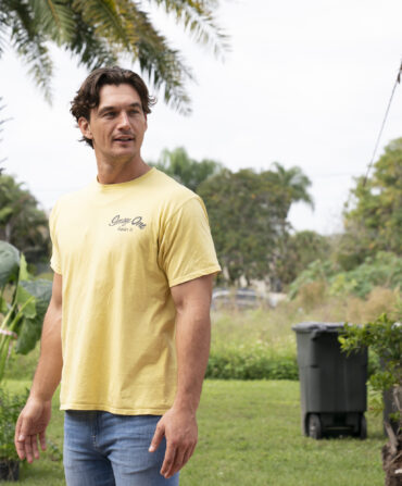 Former Bachelorette star Tyler Cameron stands in a landscape with palm trees.