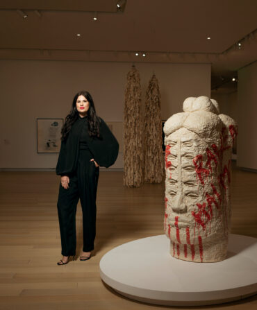 A woman in a black suit stands in a gallery next to a totemic head sculpture with many eyes and red glaze that says "Raven Halfmoon" on the side