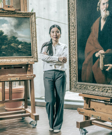 A woman stands in an art conservation studio with a large painting on an easel behind her