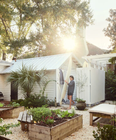 A woman holds a surfboard in a yard of a cottage with wood planting beds