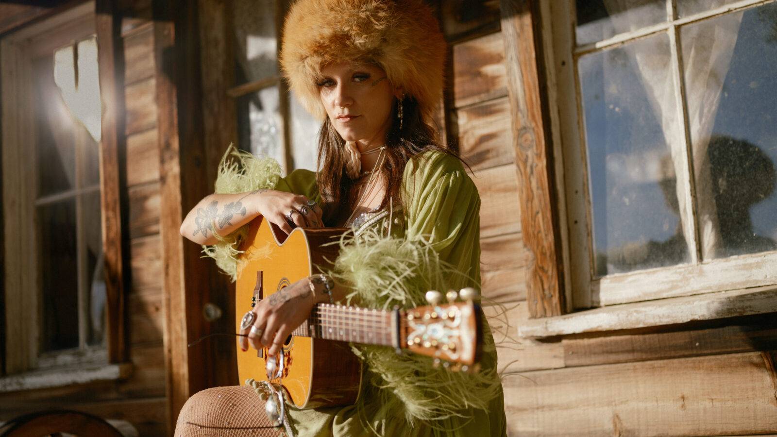 A woman, wearing a fuzzy hat and a green feathery robe, holds a guitar on a wood porch