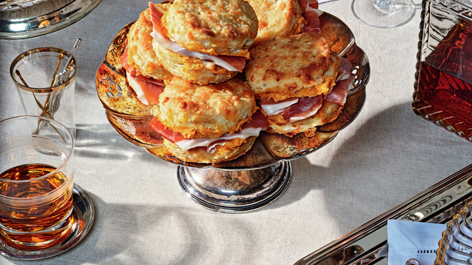 A stack of biscuits on a silver tray next to a bowl of olives and glass of bourbon