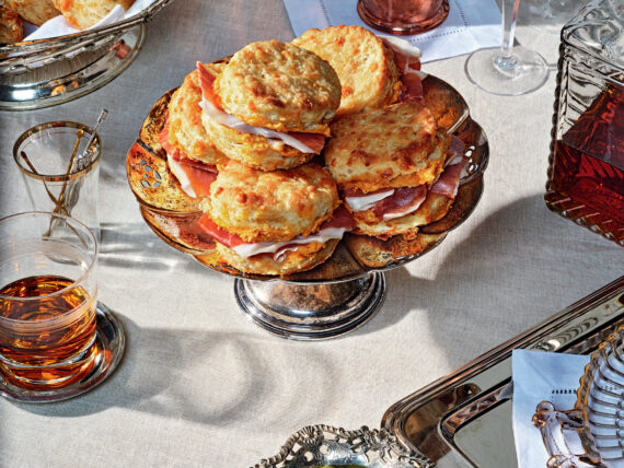 A stack of biscuits on a silver tray next to a bowl of olives and glass of bourbon