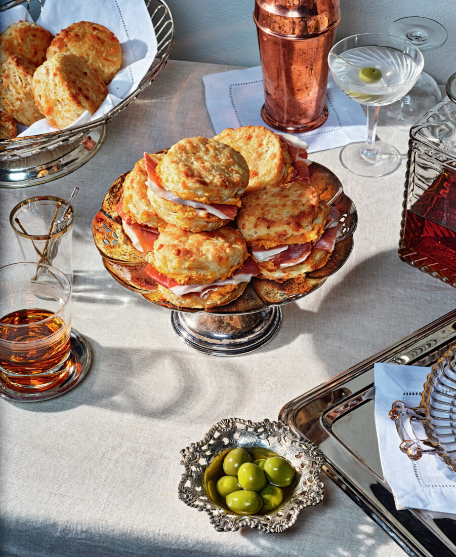 A stack of biscuits on a silver tray next to a bowl of olives and glass of bourbon