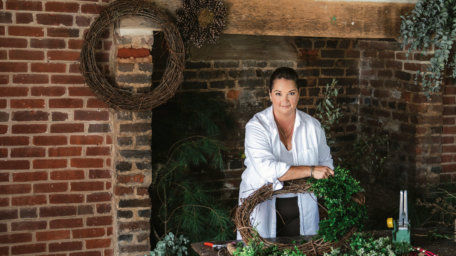 A woman stands behind a table with wreath-making supplies. There is a Spinone Italiano dog at her feet. A giant brick fireplace is behind her, and wicker baskets hang from the ceiling.