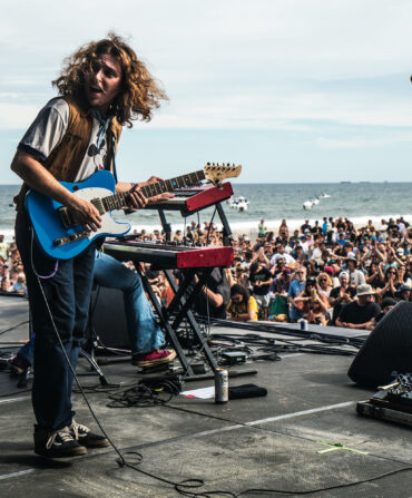A guitar player stands on a stage with a blue guitar and a microphone, turning his head back; a crowd on the beach gathers at the stage