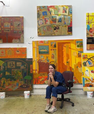 A woman sits in an art studio surrounded by colorful large paintings