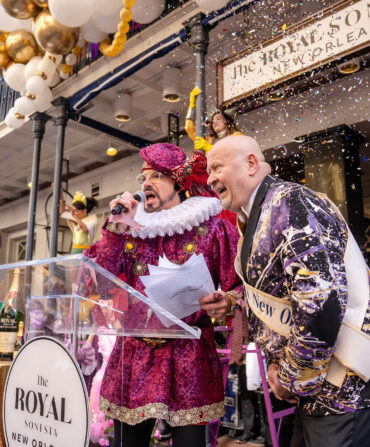 Two men dressed up for Mardi Gras stand in front of poles by a building with gold and white balloons