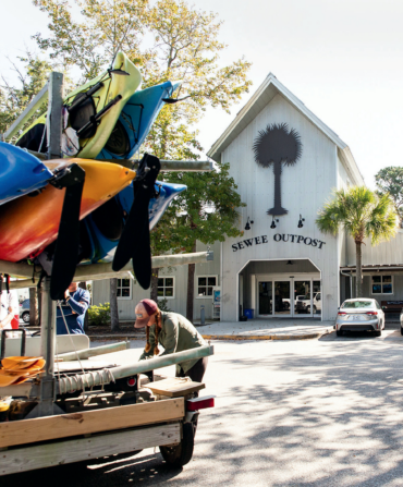 A woman stands over a car with kayaks in front of a parking lot to the Sewee Outpost